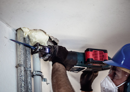 Person med blå hjälm och ansiktsmask använder en elektrisk såg för att kapa ett metallrör nära en vägg.