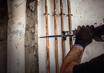 En person i handskar skär ett rör med en såg bredvid flera kopparrör på en vägg.