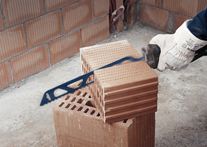 En person med handskar håller en såg mot en hög rödbruna byggstenar på en byggarbetsplats.