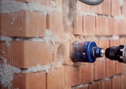 Borrmaskin med cirkelsåg borrar i röd tegelvägg, damm syns runt öppningen.