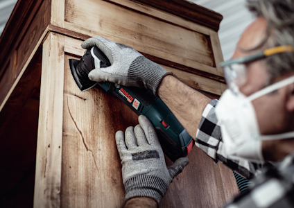 En person slipar en trämöbel med en Bosch-slipmaskin, bärandes handskar och ansiktsmask.