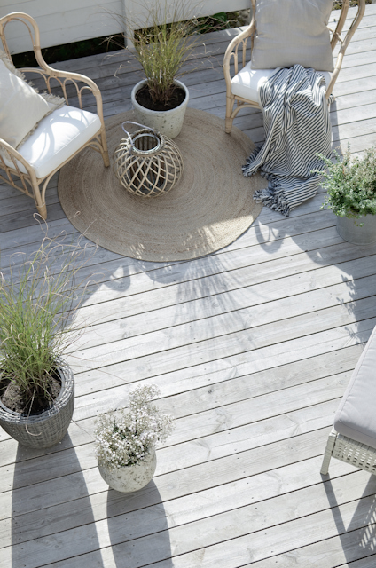 Trämöbler med dynor och kuddar på en träterrass med krukväxter och en rund matta.