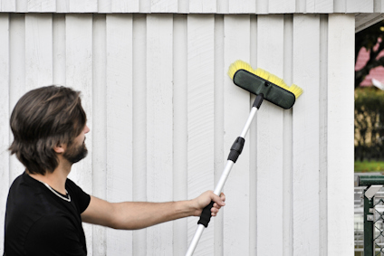 En person tvättar en vit trävägg med en borste på ett långt skaft.
