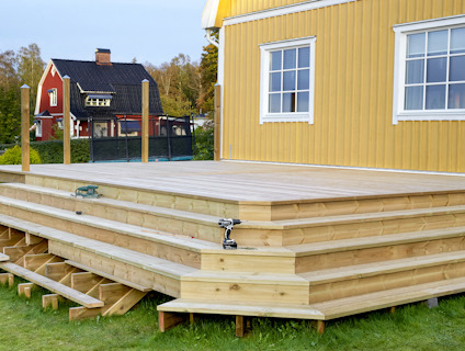 Trädäck byggt med trätrall, intill gult hus med vita fönster och röd byggnad i bakgrunden.