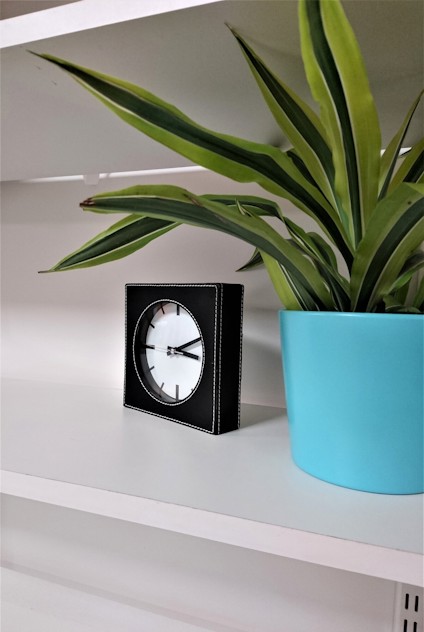 Växt i turkos kruka bredvid en kvadratisk klocka med svart ram på vit hylla.