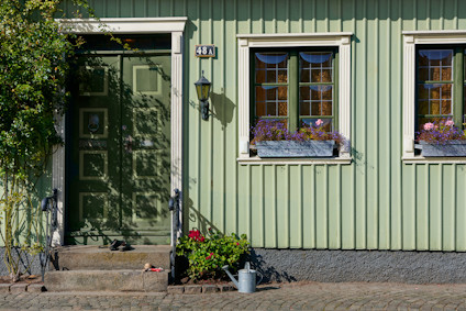 Grönt hus med två fönster med blomsterlådor, grön dörr och trappa. Växter bredvid dörren, vattenkanna på marken.