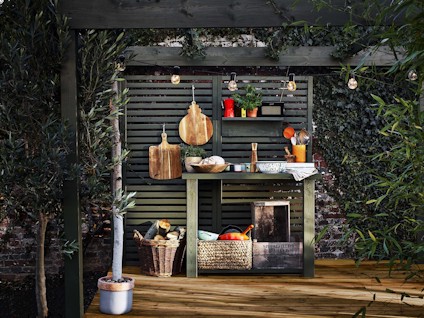 Utekök med arbetsbänk, grön spaljé, hängande träskärbrädor, korgar och redskap under glödlampor, omgiven av växter.