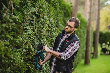 Man klipper häck med batteridriven häcksax, iklädd rutskjorta, väst och skyddsglasögon, omgiven av träd och gräsmatta.
