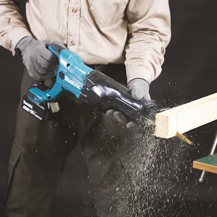 Person i arbetskläder sågar träbit med batteridriven motorsåg, sågspån flyger.