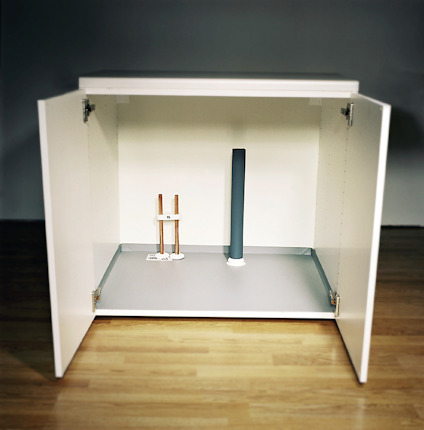 Skåp med öppna dörrar, innehållande två synliga rör och en vit basplatta. Rummet har ett trägolv.