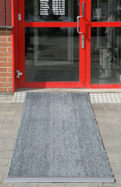 Grå dörrmatta på plattor framför röd glasdörr med öppettider.