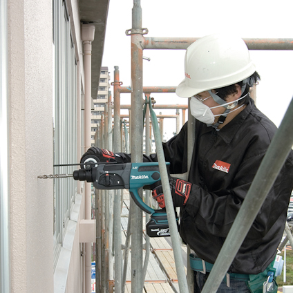 En person i skyddshjälm och jacka använder en Makita-borrmaskin vid ett fönster, omgiven av byggnadsställningar.