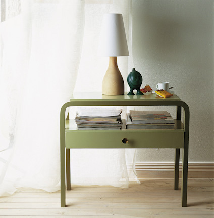 Grönt sidobord med hylla och låda, dekorerat med en lampa, vas, och tidningar, framför ett fönster med gardin.