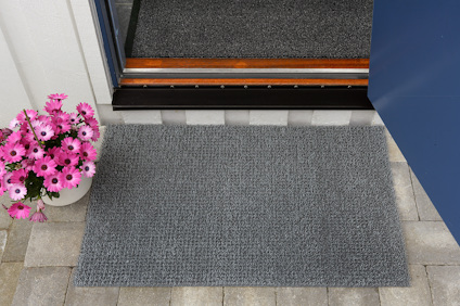 Grå dörrmatta på stenplattor, med rosa blommor i kruka till vänster.