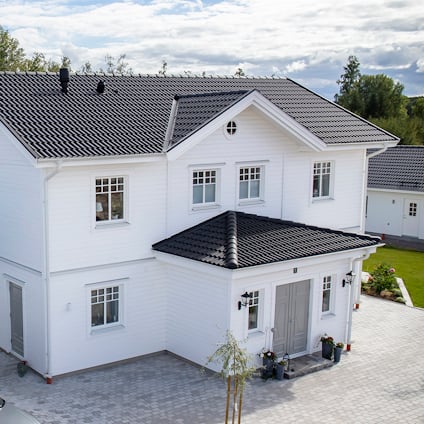 Tvåvånings hus med vita träfasader, svarta takpannor och en stensatt uppfart. Omgivande grönska och molnig himmel.