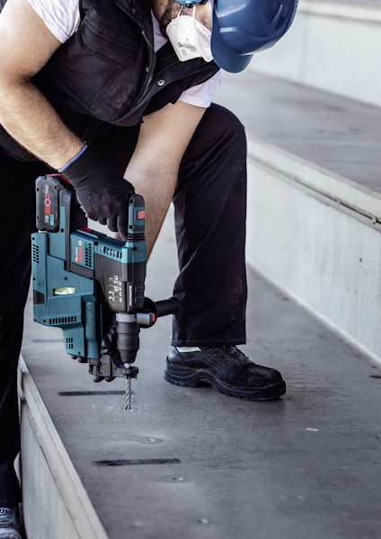 Person använder en blå och svart borrmaskin för att borra i betongtrappa, bär skyddsmask.
