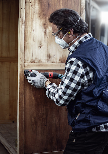 Person som använder en oscillerande multiverktyg på en träyta, iförd skyddsutrustning som glasögon och munskydd.