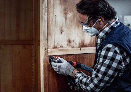 Person med skyddsglasögon, mask och handskar slipar en träpanel med en elektrisk slipmaskin.