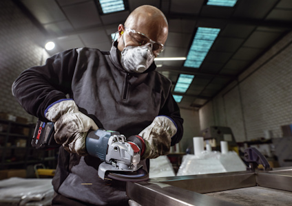 En person slipar metall med en vinkelslip i en industrimiljö, bär skyddsglasögon och ansiktsmask.