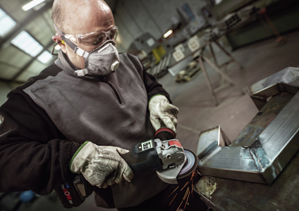 Person i skyddsmask och handskar slipar metallbit med en vinkelslip i verkstad.