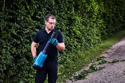 Person i skyddsglasögon använder en lövblås vid en grusväg och häck.