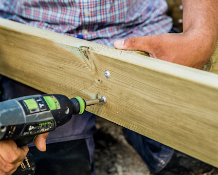 En person i färgglad skjorta använder en borrmaskin för att fästa en skruv i en träplanka.