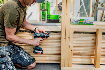 En person håller en skruvdragare och skruvar fast träreglar. Skruvar och verktyg syns i bakgrunden.