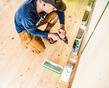 En person i blå tröja och bruna byxor skruvar i ett trägolv med en elektrisk skruvdragare. På golvet står en ask med långa konstruktionsskruvar.