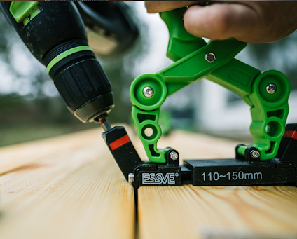 En person använder en skruvdragare och ett grönt monteringsverktyg för att skruva fast trallbrädor med jämnt mellanrum.