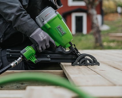 En person använder en tryckluftsdriven grön och svart spikpistol för att fästa trall utomhus.
