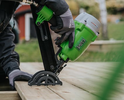 En person med arbetshandskar använder en grön och svart spikpistol för att spika fast trall vid ett altanbygge utomhus.