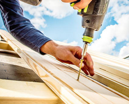 En person använder en skruvdragare för att fästa en träskruv med försänkt huvud i träplankor utomhus under en klarblå himmel.