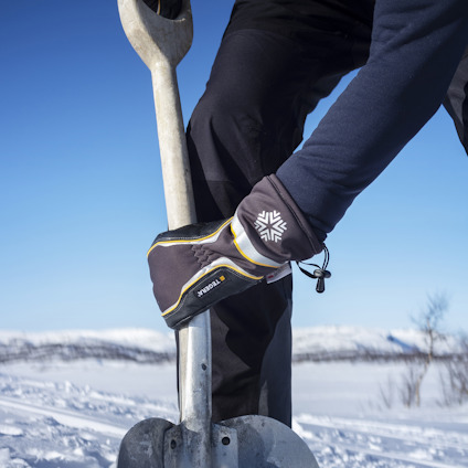 Person i vinterkläder håller en snöskyffel i en snöig landskap med träd i bakgrunden.
