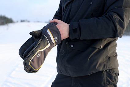Person klädd i svart jacka håller svarta vantar med gul kant och vitt motiv. Snöig bakgrund.