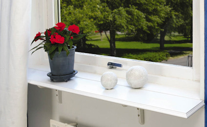 Fönsterbräda med röd blomma i kruka och två dekorativa vita bollar. Utsikt mot grön trädgård utanför.