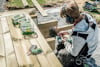 Person bygger med träplankor utomhus, med skruv och borrmaskin. lövträ produkter och skyddsutrustning syns.