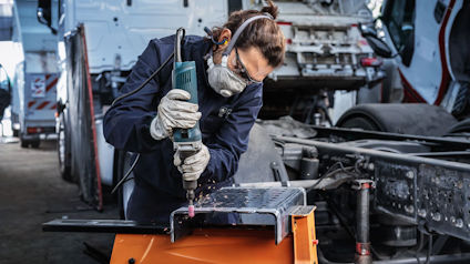 En person slipar en metallbit med en elslipmaskin. Personen bär skyddsglasögon och andningsmask.