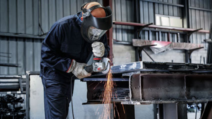 Person i skyddsutrustning slipar metall i verkstad, gnistor flyger från slipmaskinen.