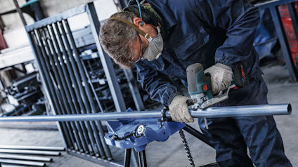 Person slipar metallrör med vinkelslip, bär skyddsglasögon och munskydd. Fler rör syns i bakgrunden.