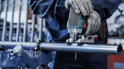 Person i skyddshandskar använder en elektrisk såg för att skära ett metallrör med hjälp av en rörklämma.
