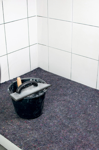 En svart hink med en spackelspade står på en filt vid ett hörn av vitt kaklat vägg.
