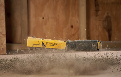 Gul och svart kniv med texten "STANLEY" och "10-800" i suddig miljö med träbakgrund och dammpartiklar i luften.