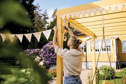 Person hänger upp vimplar på en gul pergola i en trädgård, hus och träd i bakgrunden.